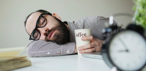 Man sleeps in office on table over laptop with coffee in hand
